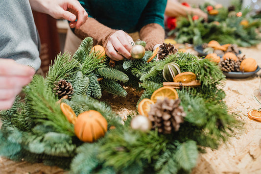 Two pairs of hands making a Christmas wreath using conifer branches, pine cones and orange slices.
