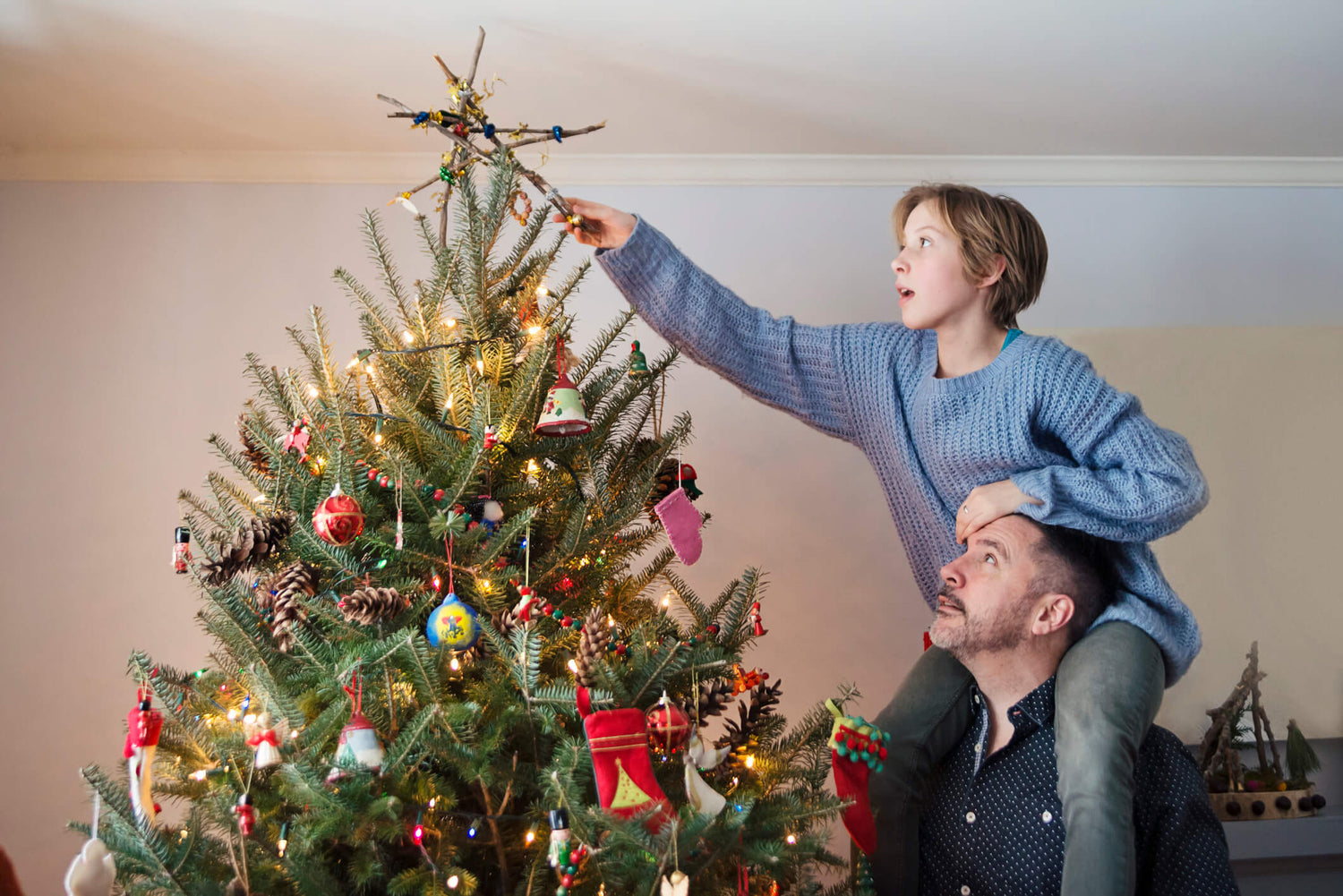 A woman, sat on a man's shoulders, places a star on top of a decorated Christmas tree