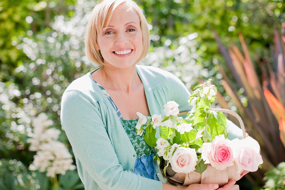 Woman with short blonde hair wearing an aqua cardigan stands in a garden holding a basket of freshly-cut pink roses.
