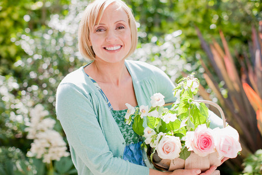Woman with short blonde hair wearing an aqua cardigan stands in a garden holding a basket of freshly-cut pink roses.