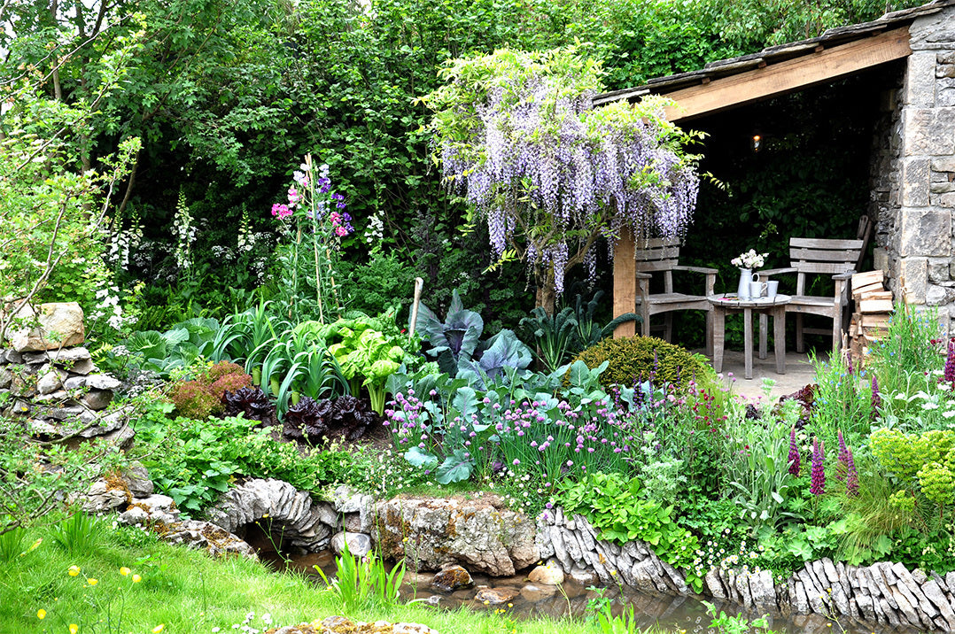 A small English cottage garden style space with a stream running through it. Behind the stream is a bed with plants and flowers and behind that a roof-topped patio area with a wisteria.